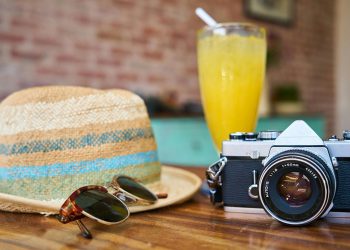 A straw hat, sunglasses, vintage camera, and a glass of orange juice on a wooden table.