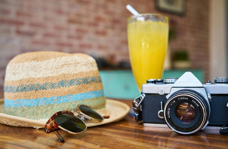 A straw hat, sunglasses, vintage camera, and a glass of orange juice on a wooden table.